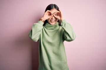 Young beautiful asian woman wearing green winter sweater over pink solated background doing ok gesture like binoculars sticking tongue out, eyes looking through fingers. Crazy expression.