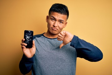 Young handsome latin man holding broken smartphone showing craked screen with angry face, negative...
