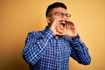 Young handsome latin man wearing casual shirt and glasses over yellow background Shouting angry out loud with hands over mouth