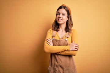 Obraz premium Young beautiful blonde girl wearing overall standing over yellow isolated background happy face smiling with crossed arms looking at the camera. Positive person.