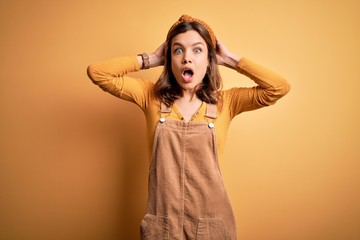 Fototapeta premium Young beautiful blonde girl wearing overall standing over yellow isolated background Crazy and scared with hands on head, afraid and surprised of shock with open mouth