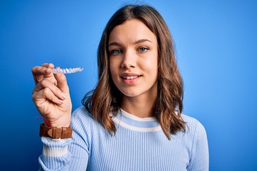 Young blonde girl holding dental orthodontic clear aligner over blue isolated background with a happy face standing and smiling with a confident smile showing teeth
