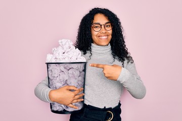 Young african american woman wearing glasses holding wastebasket with cumpled papers very happy pointing with hand and finger