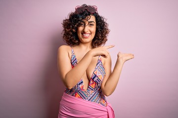 Young beautiful arab woman on vacation wearing swimsuit and sunglasses over pink background amazed and smiling to the camera while presenting with hand and pointing with finger.