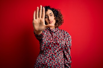 Young beautiful curly arab woman wearing casual floral dress standing over red background doing stop sing with palm of the hand. Warning expression with negative and serious gesture on the face.