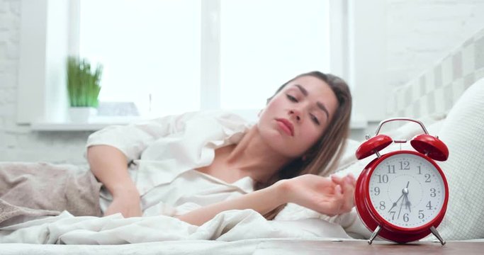 Beautiful sleepy girl lying in bed, turning off the alarm Clock and covering with Blanket in the Morning.Young Woman continues to sleep after Alam Clock, Lazy Morning. Home Quarantine. Staying Indoor.