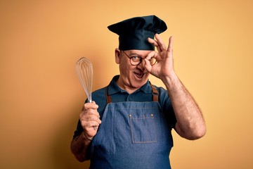 Middle age hoary cooker man wearing apron and hat holding whisk over yellow background with happy face smiling doing ok sign with hand on eye looking through fingers