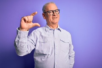 Middle age handsome hoary man wearing striped shirt and glasses over purple background smiling and confident gesturing with hand doing small size sign with fingers looking and the camera. Measure