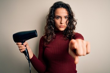 Young beautiful woman with curly hair using hair dryer over isolated white background pointing with finger to the camera and to you, hand sign, positive and confident gesture from the front