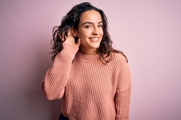 Obraz premium Young beautiful woman with curly hair wearing casual sweater over isolated pink background smiling with hand over ear listening an hearing to rumor or gossip. Deafness concept.