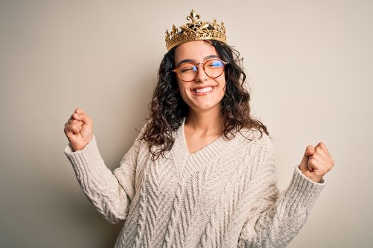 Young beautiful woman with curly hair wearing golden queen crown over white background very happy and excited doing winner gesture with arms raised, smiling and screaming for success. Celebration