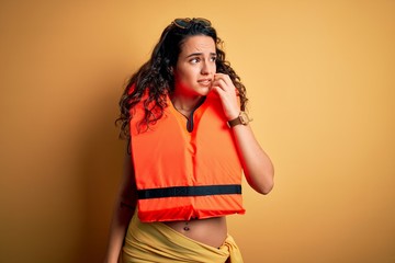 Young beautiful woman with curly hair wearing orange lifejacket over yellow background looking...