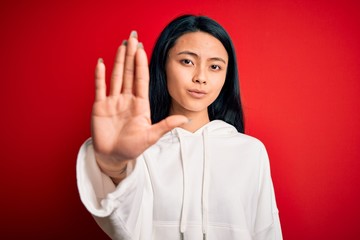 Young beautiful chinese sporty woman wearing sweatshirt over isolated red background doing stop sing with palm of the hand. Warning expression with negative and serious gesture on the face.