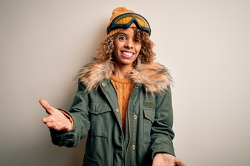 Young african american skier woman with curly hair wearing snow sportswear and ski goggles smiling cheerful with open arms as friendly welcome, positive and confident greetings