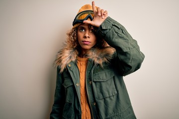 Young african american skier woman with curly hair wearing snow sportswear and ski goggles making fun of people with fingers on forehead doing loser gesture mocking and insulting.