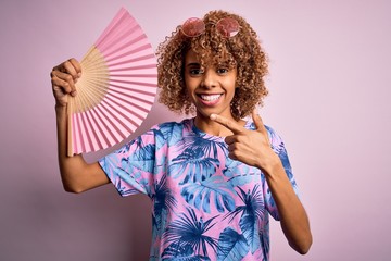 Young african american curly woman on vacation using hand fan over isolated pink background very happy pointing with hand and finger