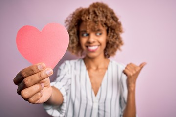 Young african american romantic woman with curly hair holding paper heart shape pointing and showing with thumb up to the side with happy face smiling