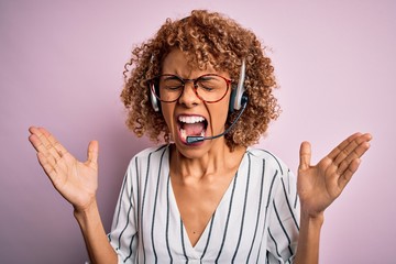 African american curly call center agent woman working using headset over pink background celebrating mad and crazy for success with arms raised and closed eyes screaming excited. Winner concept