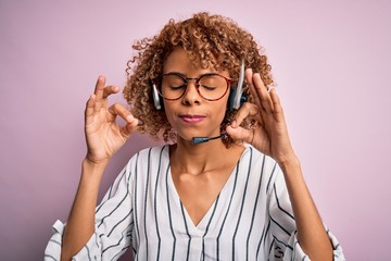 African american curly call center agent woman working using headset over pink background relax and smiling with eyes closed doing meditation gesture with fingers. Yoga concept.