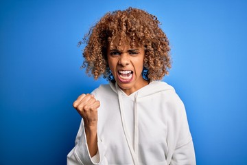 Fototapeta na wymiar Beautiful african american sporty woman wearing casual sweatshirt over blue background angry and mad raising fist frustrated and furious while shouting with anger. Rage and aggressive concept.