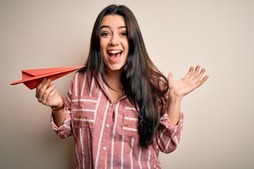 Young brunette woman holding origami paper plane over isolated background very happy and excited, winner expression celebrating victory screaming with big smile and raised hands