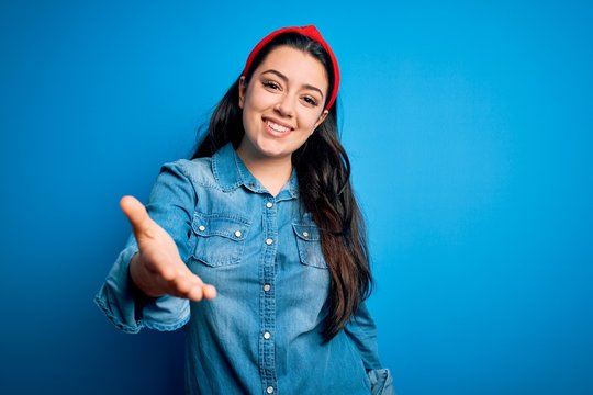 Young brunette woman wearing casual denim shirt over blue isolated background smiling friendly offering handshake as greeting and welcoming. Successful business.