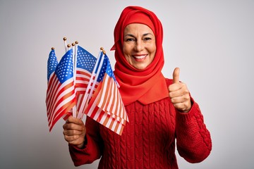Middle age patriotic woman wearing traditional muslim hijab holding united states flags happy with big smile doing ok sign, thumb up with fingers, excellent sign