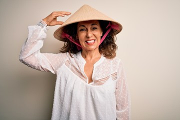 Middle age brunette woman wearing asian traditional conical hat over white background confuse and wonder about question. Uncertain with doubt, thinking with hand on head. Pensive concept.