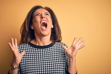 Middle age beautiful woman wearing casual sweater over isolated yellow background crazy and mad shouting and yelling with aggressive expression and arms raised. Frustration concept.