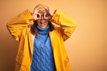 Middle age woman wearing yellow raincoat and winter hat over isolated background doing ok gesture like binoculars sticking tongue out, eyes looking through fingers. Crazy expression.