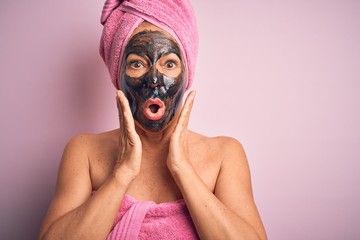 Middle age brunette woman wearing beauty black face mask over isolated pink background afraid and shocked, surprise and amazed expression with hands on face