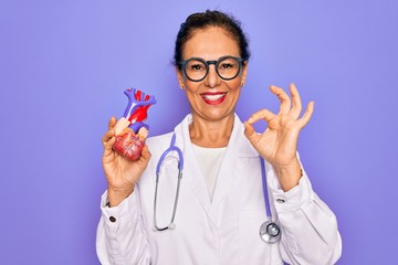 Middle age senior cardiologist doctor woman holding professional cardiology heart doing ok sign with fingers, excellent symbol - obrazy, fototapety, plakaty
