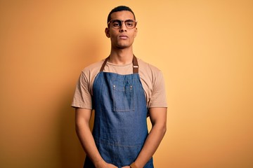 Young handsome african american shopkeeper man wearing apron over yellow background with serious expression on face. Simple and natural looking at the camera.