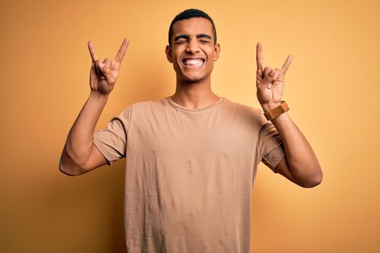Young handsome african american man wearing casual t-shirt standing over yellow background shouting with crazy expression doing rock symbol with hands up. Music star. Heavy music concept.