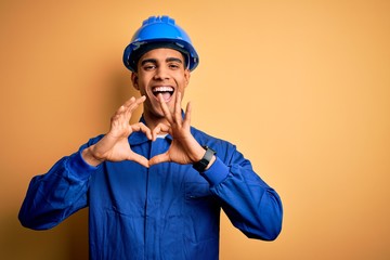 Young handsome african american worker man wearing blue uniform and security helmet smiling in love showing heart symbol and shape with hands. Romantic concept.