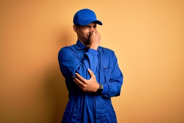 Young african american mechanic man wearing blue uniform and cap over yellow background smelling...