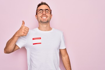 Handsome man with blue eyes wearing sticker with hello my name is message identification doing happy thumbs up gesture with hand. Approving expression looking at the camera showing success.