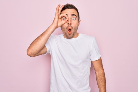 Handsome man with blue eyes wearing casual white t-shirt standing over pink background doing ok gesture shocked with surprised face, eye looking through fingers. Unbelieving expression.