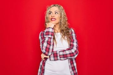 Young beautiful blonde woman wearing casual shirt standing over isolated red background with hand on chin thinking about question, pensive expression. Smiling and thoughtful face. Doubt concept.