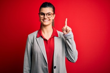 Young beautiful brunette businesswoman wearing jacket and glasses over red background showing and pointing up with finger number one while smiling confident and happy.