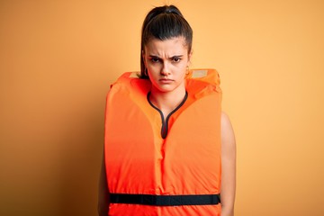 Young beautiful brunette woman wearing orange safe lifejacket over yellow background skeptic and...