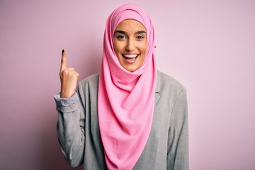 Young beautiful brunette businesswoman wearing pink muslim hijab and business jacket pointing finger up with successful idea. Exited and happy. Number one.