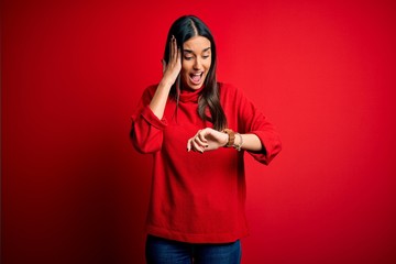 Young beautiful brunette woman wearing casual sweater over isolated red background Looking at the watch time worried, afraid of getting late