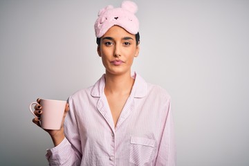 Young beautiful brunette woman wearing pajama and sleep mask drinking cup of coffee with a confident expression on smart face thinking serious