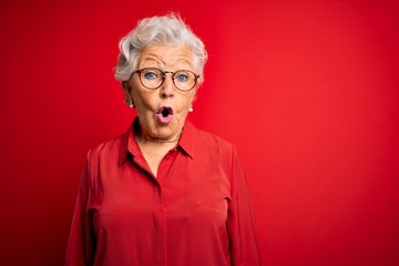 Senior beautiful grey-haired woman wearing casual shirt and glasses over red background afraid and shocked with surprise expression, fear and excited face. - obrazy, fototapety, plakaty