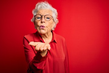 Senior beautiful grey-haired woman wearing casual shirt and glasses over red background looking at the camera blowing a kiss with hand on air being lovely and sexy. Love expression. - obrazy, fototapety, plakaty