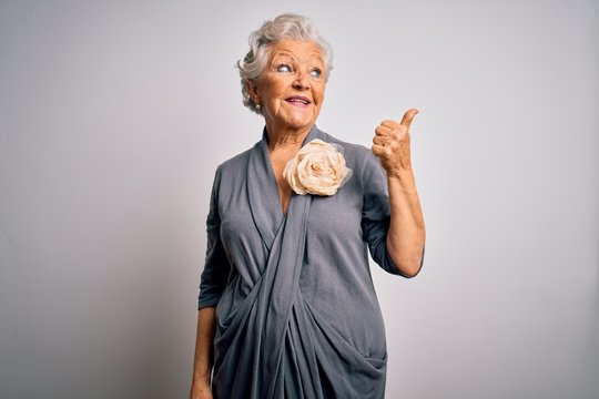 Senior beautiful grey-haired woman wearing casual dress standing over white background smiling with happy face looking and pointing to the side with thumb up.