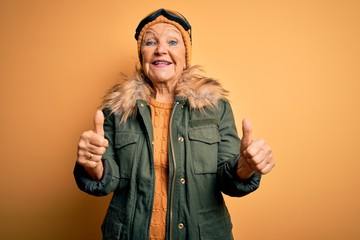 Senior beautiful grey-haired skier woman wearing snow sportswear and ski goggles approving doing positive gesture with hand, thumbs up smiling and happy for success. Winner gesture.