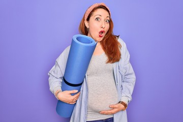 Young beautiful redhead pregnant woman expecting baby holding mat to do prenatal exercise scared in shock with a surprise face, afraid and excited with fear expression