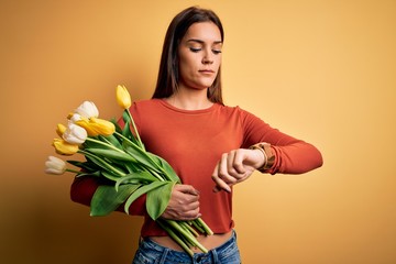 Young beautiful brunette woman holding bouquet of tulips flowers over yellow background Checking the time on wrist watch, relaxed and confident